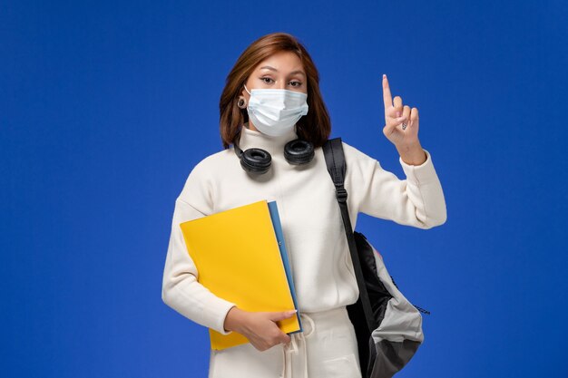 Front view young female student in white jersey wearing mask and backpack holding files on the blue wall