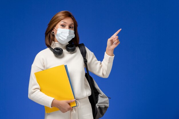 Front view young female student in white jersey wearing mask and backpack holding files on the blue wall