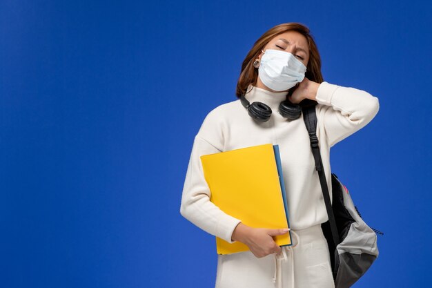 Front view young female student in white jersey wearing mask and backpack holding files having neckache on blue wall