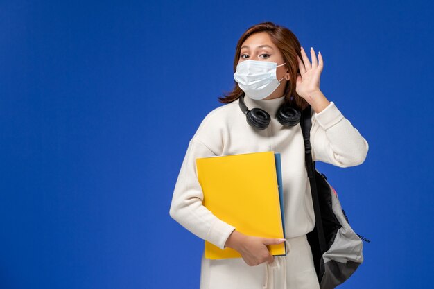 Front view young female student in white jersey wearing mask and backpack holding files trying to hear on blue wall