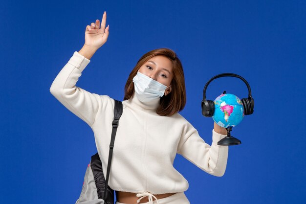 Вид спереди молодая студентка в белой майке в маске и рюкзаке, держащая глобус с наушниками на синей стене