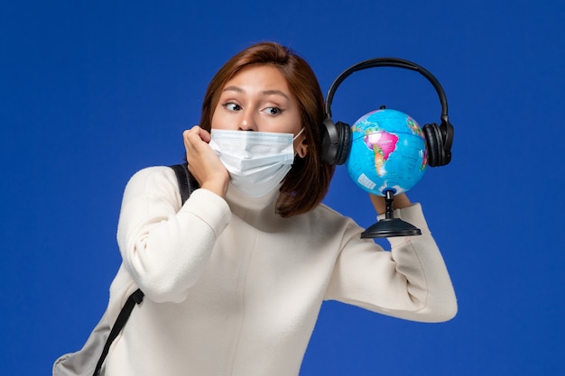 Free photo front view young female student in white jersey wearing mask and backpack holding globe with headphones on blue wall