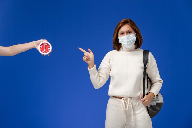 Front view young female student in white jersey wearing mask and bag pointing out at clock on blue wall