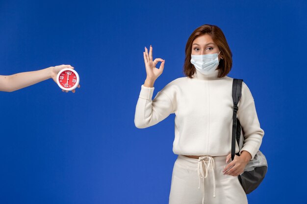 Молодая студентка в белом свитере в маске и сумке, вид спереди, показывает знак на синей стене