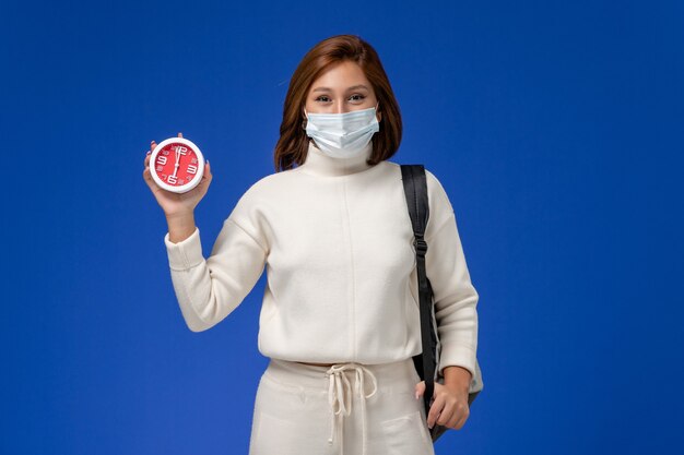 Front view young female student in white jersey wearing mask and holding clock on blue wall