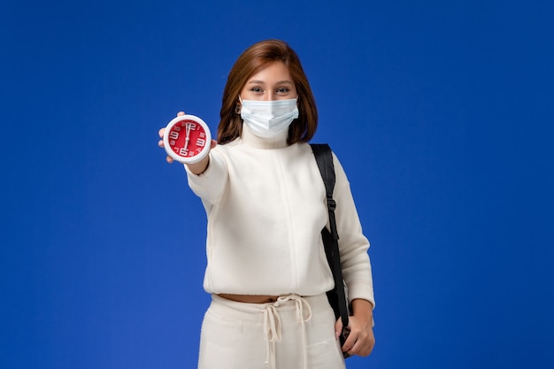 Vista frontale giovane studentessa in maglia bianca che indossa la maschera e tenendo l'orologio sulla parete blu