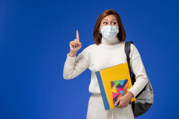 青い壁にバッグとコピーブックとマスクを身に着けている白いジャージの正面図若い女子学生