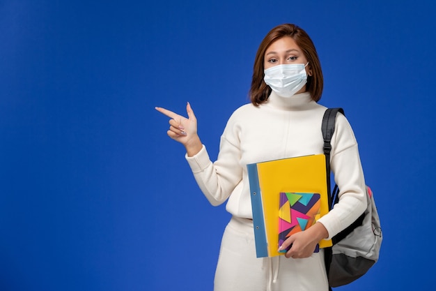 Front view young female student in white jersey wearing mask with bag and copybooks on the blue wall