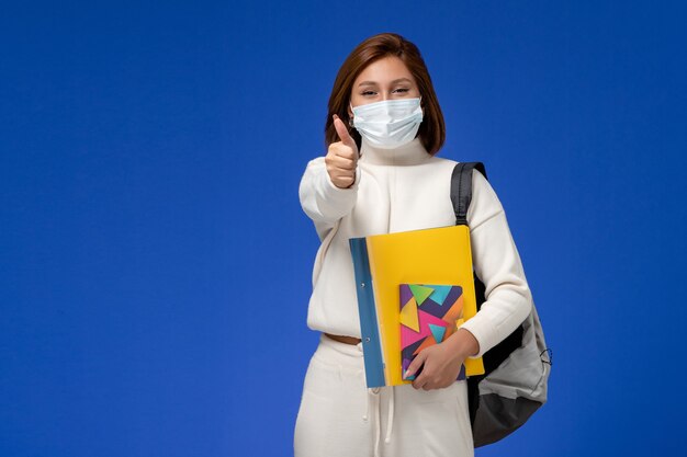 Вид спереди молодая студентка в белом джерси в маске с сумкой и тетрадями на синей стене