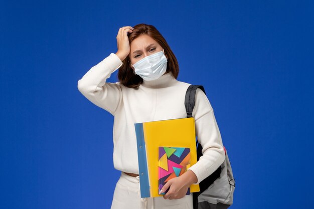 Front view young female student in white jersey wearing mask with bag and copybooks having headache on the blue wall