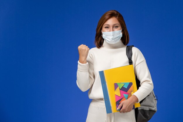 Front view young female student in white jersey wearing mask with bag and copybooks rejoicing on the blue wall