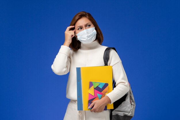 Front view young female student in white jersey wearing mask with bag and copybooks thinking on blue wall