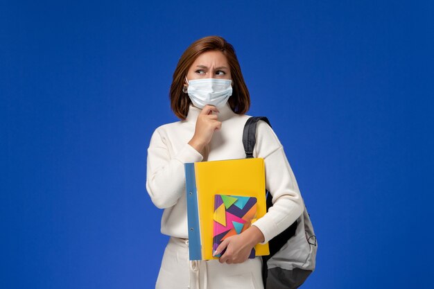 Front view young female student in white jersey wearing mask with bag and copybooks thinking on the blue wall