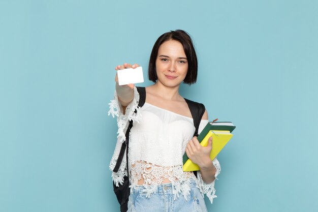Front view young female student in white shirt blue jeans and black bag holding copybooks and card on the blue space female student 