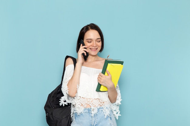 Front view young female student in white shirt blue jeans and black bag holding copybooks talking on the phone on the blue space female student university teach