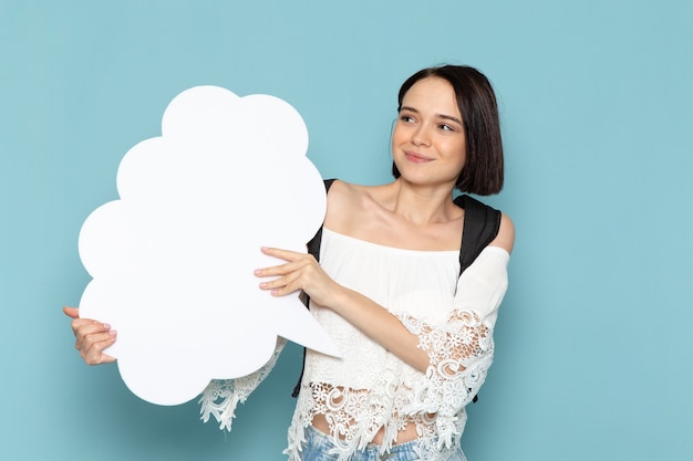 Front view young female student in white shirt blue jeans and black bag holding enormous white sign smiling on the blue space female student university