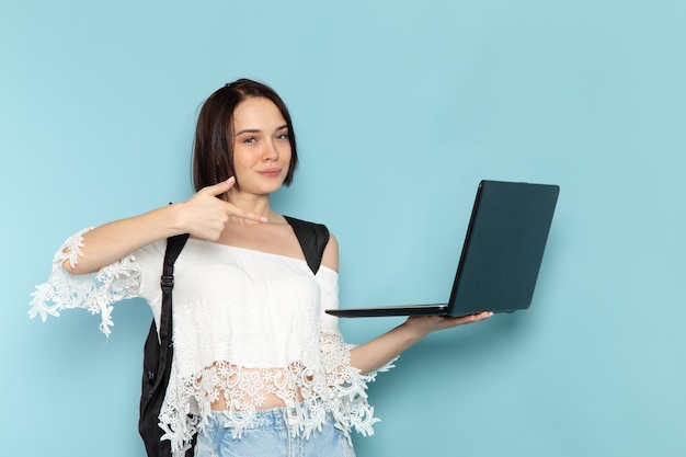 Front view young female student in white shirt blue jeans and black bag using laptop on the blue space female student university school