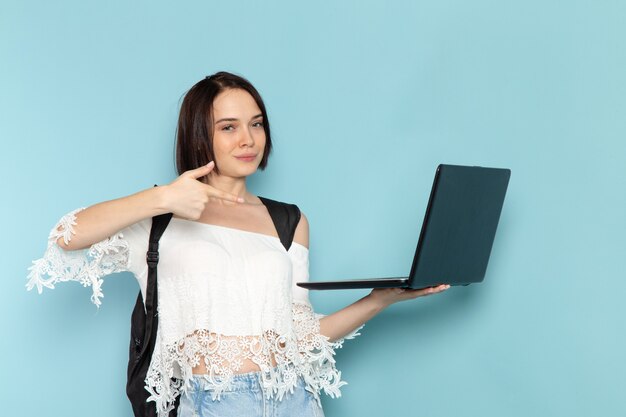 Front view young female student in white shirt blue jeans and black bag using laptop on the blue space female student university school