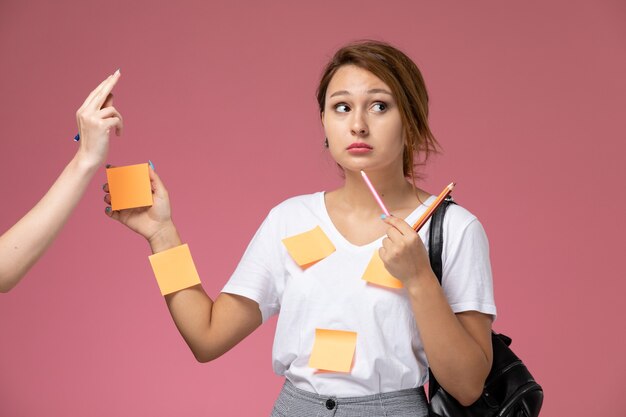 Front view young female student in white t-shirt and grey trousers posing with pencils and papers on pink background student lessons university college