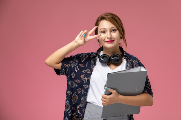 Giovane studentessa di vista frontale in maglietta bianca e pantaloni grigi che sorride con gli auricolari sull'istituto universitario di lezioni dello studente di sfondo rosa