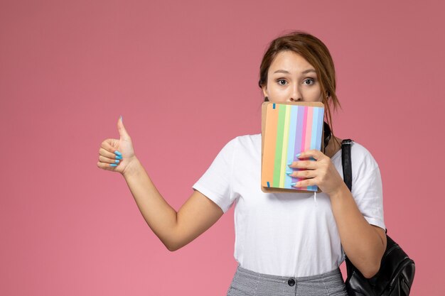 ピンクの背景のレッスン大学大学で彼女の手にコピーブックを持つ白いTシャツと灰色のズボンの正面図若い女子学生