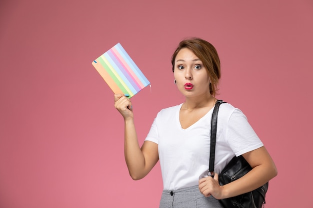 Front view young female student in white t-shirt and grey trousers with copybook in her hands on pink background student lessons university college