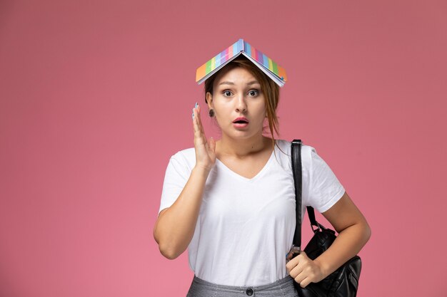 Front view young female student in white t-shirt and grey trousers with copybook on her head on pink background student lessons university college