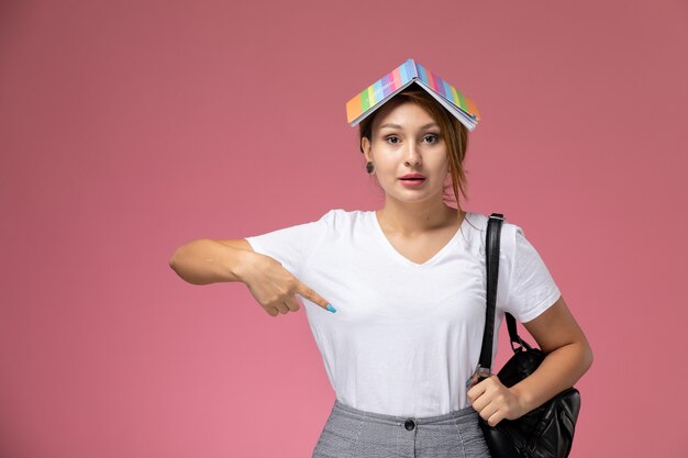 Front view young female student in white t-shirt and grey trousers with copybook on her head on pink background student lessons university college