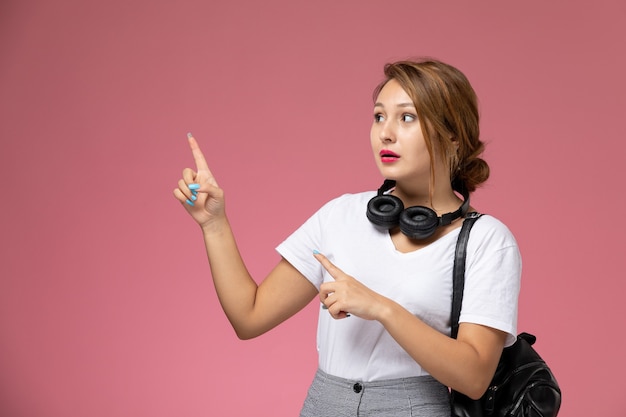 Front view young female student in white t-shirt and grey trousers with earphones posing on pink background student lessons university college