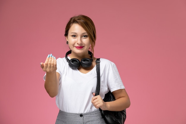 正面図白いTシャツとイヤフォンとピンクの背景の笑顔の表情で灰色のズボンの若い女子学生学生レッスン大学