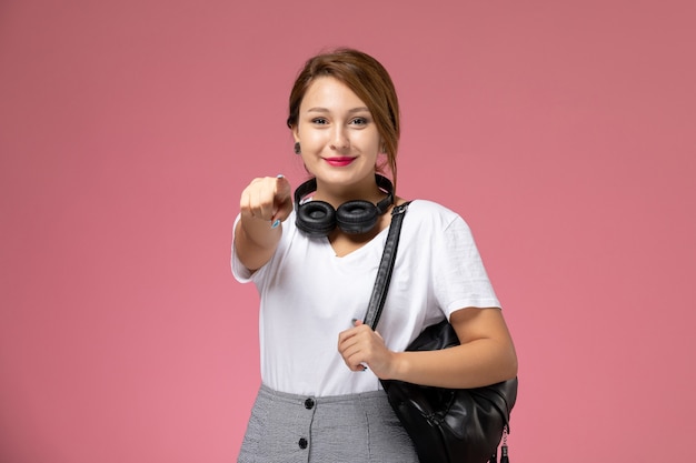 Front view young female student in white t-shirt and grey trousers with earphones smiling on pink background student lessons university college