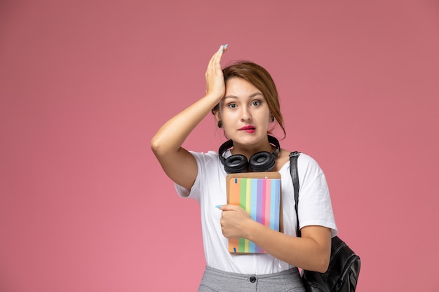 분홍색 배경 학생 수업 대학 대학에 그녀의 손에 헤드폰과 카피 북 흰색 티셔츠와 회색 바지에 전면보기 젊은 여성 학생