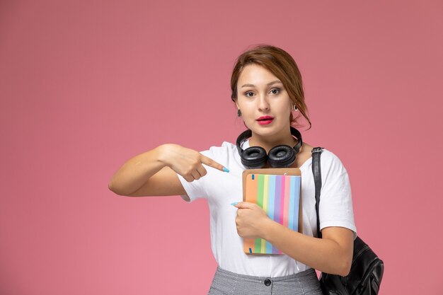 ピンクの背景のレッスン大学大学の研究書にコピーブックを保持している白いTシャツの正面図若い女子学生