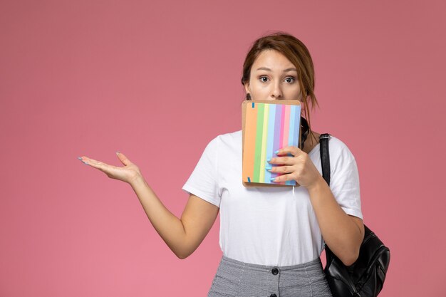 ピンクの背景に驚きの表情でコピーブックを保持している白いTシャツの正面図若い女子学生レッスン大学大学研究書