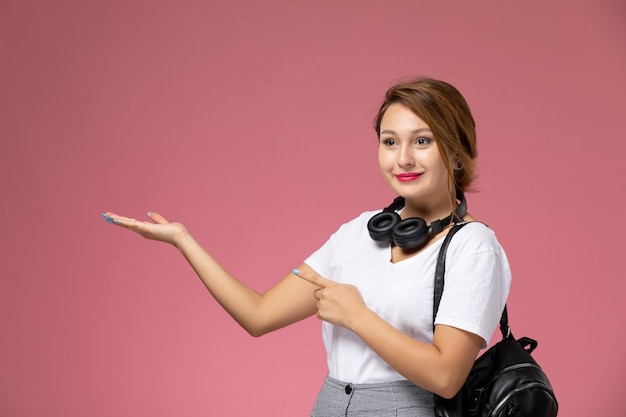 Front view young female student in white t-shirt with bag and earphones posing and smiling on pink background lesson university college study book