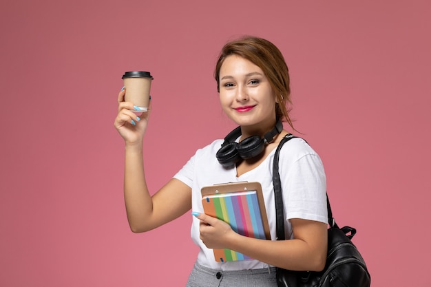 Front view young female student in white t-shirt with copybook and bag posing and holding coffee cup on pink background lesson university college study book