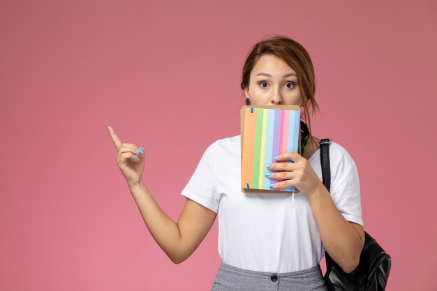 Front view young female student in white t-shirt with copybook and bag posing on pink background lesson university college study book