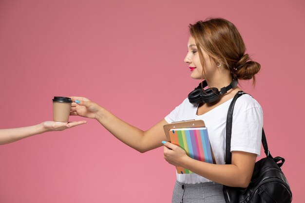 Front view young female student in white t-shirt with copybook and bag posing and taking coffee cup on the pink background lesson university college study book