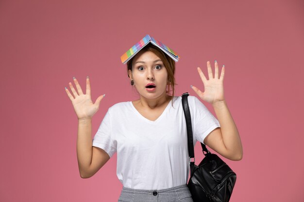 Front view young female student in white t-shirt with copybook and bag posing with shocked expression on pink background lesson university college study book