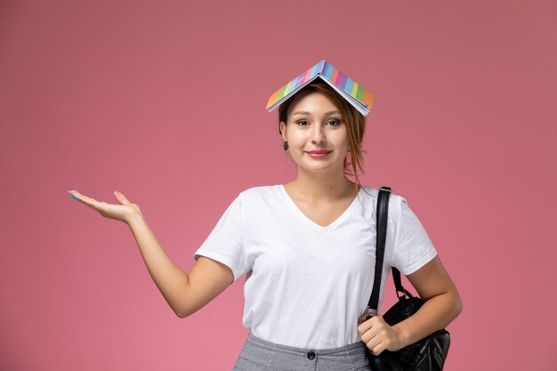 Front view young female student in white t-shirt with copybook and bag posing and with a slight smile on pink background lesson university college study book