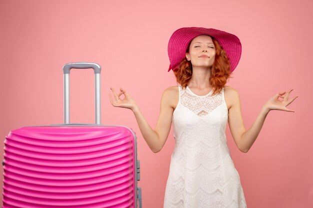 Front view of young female tourist with pink hat and bag meditating on pink wall