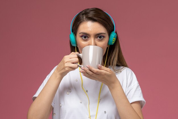 Вид спереди молодая женщина в белой рубашке пьет кофе, слушая музыку на розовой стене модельная женщина