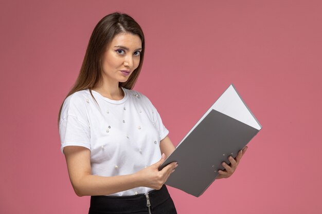 Giovane femmina di vista frontale in camicia bianca che tiene documento grigio sulla parete rosa, modello di posa della donna di colore