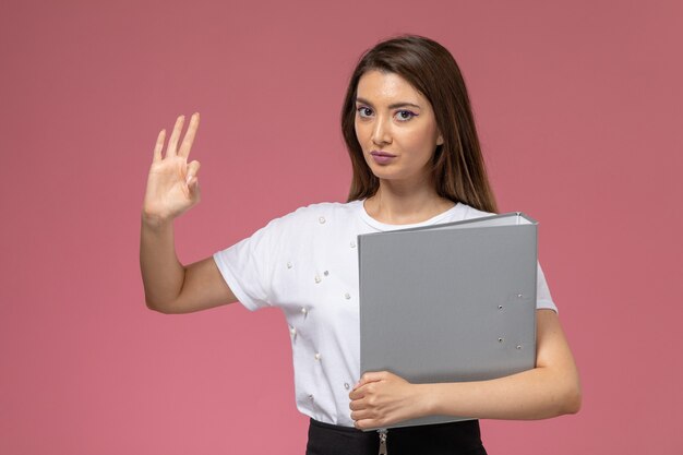 Front view young female in white shirt holding grey file on the pink wall, color woman pose model woman