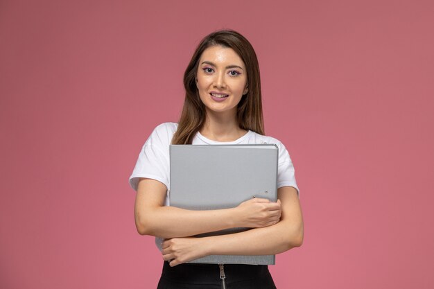 Front view young female in white shirt holding grey file with smile on the pink wall, color woman pose model woman