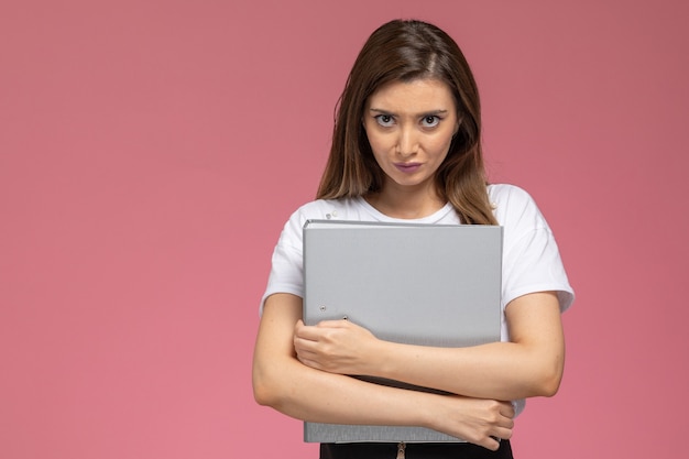 Front view young female in white shirt holding grey files on light pink wall, model woman pose