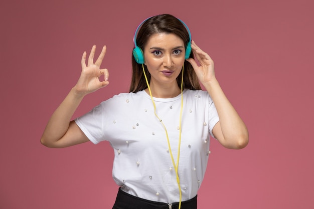 Front view young female in white shirt listening to music and posing on the pink wall, color woman model