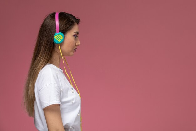 Вид спереди молодая женщина в белой рубашке, слушающая музыку в наушниках на розовой стене, цветная женщина позы модель женщина