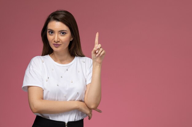 Front view young female in white shirt posing with raised finger on pink wall, color woman pose model