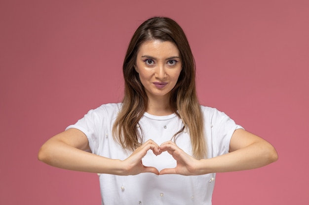 Front view young female in white shirt showing heart and love sign on the pink wall, color woman model